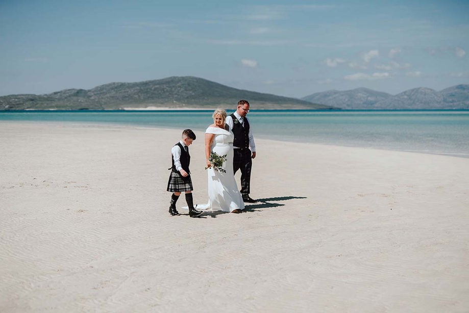 Bride and groom with child in kilt walking along a white sandy beach with hills and blue sea in the background